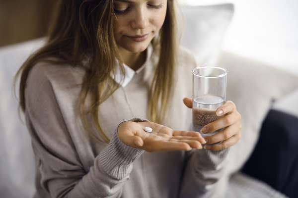 Eine braunhaarige Frau hält in ihrer linken Hand ein Glas mit Wasser und schaut auf eine Naproxentablette in der anderen Hand.