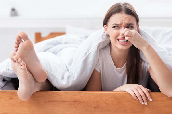 Eine Frau liegt im Bett und hält sich die Nase wegen der Füße neben ihr zu.