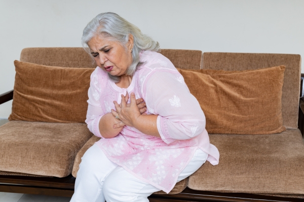 Ältere, übergewichtige Frau sitzt auf einem Sofa und hält sich ihre Herzregion vor Schmerzen