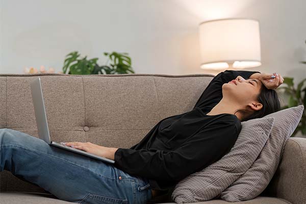 Eine frau liegt mit ihren Notebook auf der Couch und hält sich mit dem rechten Arm die Stirn Eine frau liegt mit ihren Notebook auf der Couch und hält sich mit dem rechten Arm die Stirn